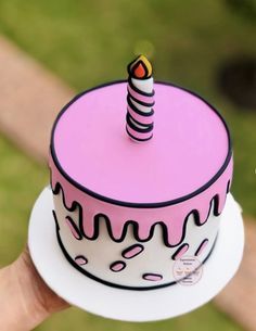 a hand holding a pink and white cake with a lit candle on it's top