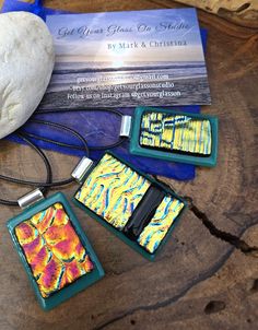 three square pendants with colorful designs on them sitting next to a rock and business card