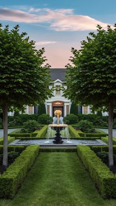 a fountain surrounded by hedges in front of a large house with trees and bushes around it