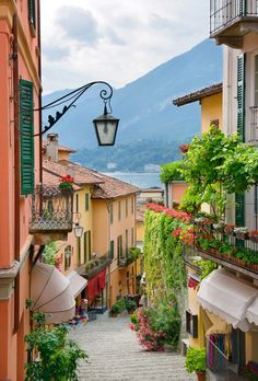 an alley way with flowers and umbrellas next to buildings on either side, in front of a mountain range