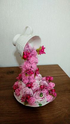 a white vase with pink flowers in it on top of a wooden table next to a wall