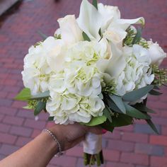 a bouquet of white flowers is held by a woman's hand on a brick sidewalk