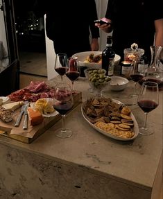 two people standing next to a table full of food and wine glasses filled with red wine