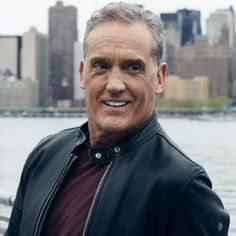 a man standing in front of a body of water wearing a black jacket and smiling