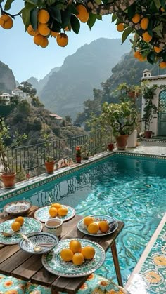 an outdoor pool with oranges and plates on the table next to it in front of mountains