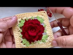 two hands holding crocheted square with a red rose on it and another person using a needle