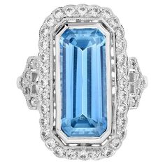 Drawing inspiration from the pattern of the bygone ear, this Art Deco-inspired blue topaz ring is crafted in 18k white gold. Glimmering round diamonds create a brilliant halo around the emerald-cut blue topaz. Additional diamond accents embellish the shoulder for extra shimmer. Ring Information Style: Art-deco Metal: 18K White Gold Weight: 5.18 g. (approx. total weight) Center Gemstones Type: Blue Topaz Shape: Emerald Cut Size: 1696 mm. Number: 1 Weight: 7 Carat (approx. total weight) Accent Gemstones Type: Diamond Shape: Round Average Color: H Average Clarity: SI Size: 1.3 – 1.6 mm. Number: 42 Weight: 0.66 Carat (approx. total weight) This ring will be made to your order in 1-2 weeks, contact us to find out more. An attractive ring box is included.  Most of our rings can be sized to any f Natural Emerald Rings, Contemporary Engagement Rings, Emerald Cut Rings, Diamond Halo Ring, Diamond Cocktail Rings, White Gold Engagement, Halo Diamond Ring, White Gold Engagement Rings, Women Diamond