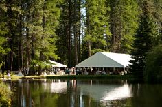 a group of people standing next to a lake under a white tent surrounded by trees