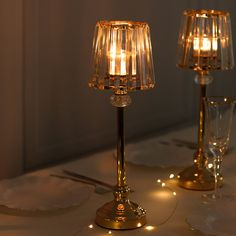 two clear glass candlesticks on a white table cloth with silverware and wine glasses