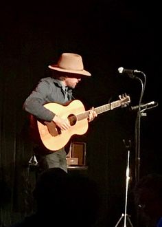 a man in a fedora playing an acoustic guitar while standing on stage with a microphone