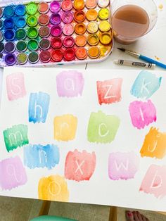 a child's hand holding up a paper with letters painted on it and watercolors in the background