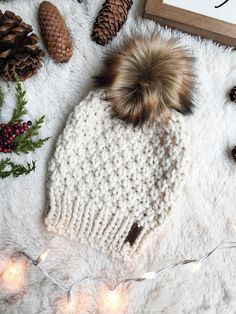 a knitted hat with a fur pom sits on a white blanket next to pine cones and christmas lights