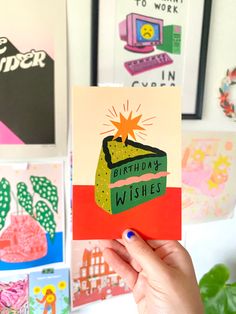 a hand holding up a birthday card in front of some art work on the wall