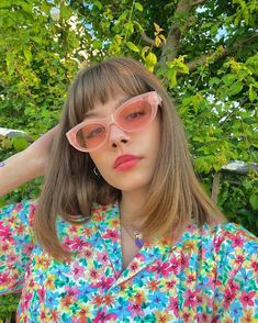 a woman wearing pink sunglasses standing in front of a green leafy tree with her hands on her head