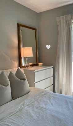 a white bed sitting under a mirror next to a lamp on a dresser in a bedroom