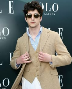 a man wearing sunglasses and a tan jacket standing in front of a green wall with his hands on his hips