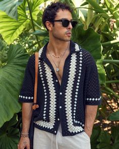 a man wearing sunglasses and a crocheted shirt stands in front of some plants