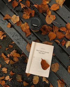 an open book sitting on top of a wooden bench covered in leaves next to a coffee cup