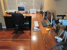 a desk with two computers and a camera in front of the window, on a hard wood floor