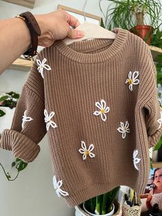 a person holding up a brown sweater with white flowers on it and a plant in the background