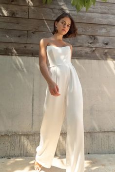 a woman standing in front of a wooden wall wearing a white jumpsuit and sandals