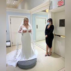 two women standing in front of a mirror wearing facemasks and veils, one woman is looking at the bride's dress