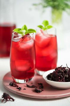 two glasses filled with red liquid next to a bowl full of blackberries and mint