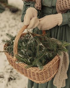 a woman holding a wicker basket filled with evergreen branches and pine cones in the snow