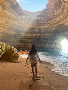 a woman is walking on the beach in front of a cave