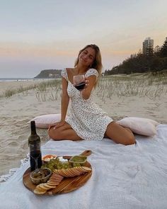 a woman sitting on the beach with a bottle of wine and plate of food next to her