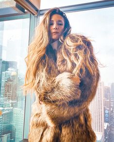 a woman standing in front of a window with long hair and wearing a fur coat