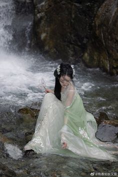 a woman in a green dress sitting on rocks next to a waterfall and holding a butterfly