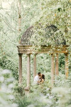 an engaged couple cuddles under a gazebo surrounded by greenery in the woods