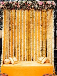 a bed with pillows and flowers on it in front of a yellow curtain that is hanging from the ceiling