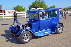 an old blue car parked in front of a white fence with people standing around it