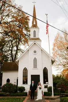 a bride and groom standing in front of a white church with an american flag on top