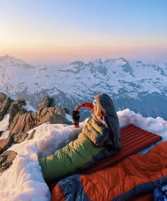 a woman sitting on top of a snow covered mountain next to an orange sleeping bag