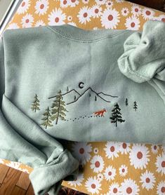 a green sweater with trees and mountains embroidered on the front, sitting on a table