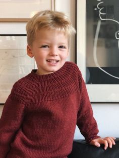 a young boy in a red sweater is sitting on a black chair and smiling at the camera