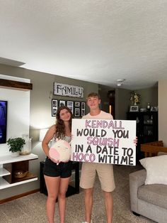 a man and woman standing in a living room holding a sign that says, rendall would you crush in a date to hoo witme?