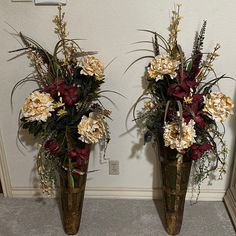 two metal vases with flowers in them on the floor next to a wall and mirror