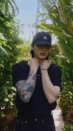 a man with tattoos on his arm and hat standing in front of some plants wearing a baseball cap