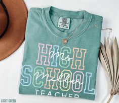 You'll adore this high school teacher shirt! Crafted by Comfort Colors, this tee combines ultimate comfort with a charming design for your high school teacher. The front features a vibrant design with "High School" in bold block letters and a stylish script font overlay of your personalized teacher's name. Available in four beautiful colors, it's the ideal gift for your child's teacher for Christmas, a birthday, or even a treat for yourself. Plus, it's a wonderful teacher appreciation gift.  Per Green School Tops With Text Print, Green Letter Print Top For School, Green Text Print T-shirt For School, Green Pre-shrunk Tops For School, Green Text Print School Tops, Green Text Print Tops For School, Green Text Print Top For School, Green Letter Print T-shirt For End Of School Year, Green Graphic Tee For School