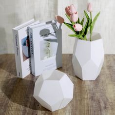 two white vases with pink flowers in them on a table next to some books