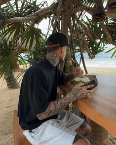 a man with tattoos sitting at a wooden table next to the ocean and holding a book