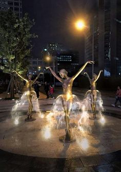 three statues in the middle of a fountain with lights shining on them and people standing around