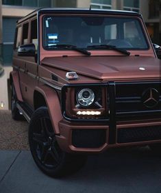 a brown mercedes g - class is parked in front of a building with its lights on