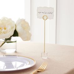 there is a place setting on the table with white flowers in a vase and gold cutlery