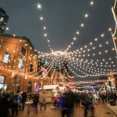 many people are walking down the street with christmas lights strung over them and buildings in the background