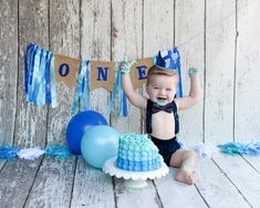 a baby boy sitting on the floor in front of a cake and balloons with blue streamers